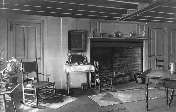 View of Cochrane House dining room. The room features a fireplace with mantle, rugs, tables and chairs, houseplants, oil lamp, candlesticks and other household items.
