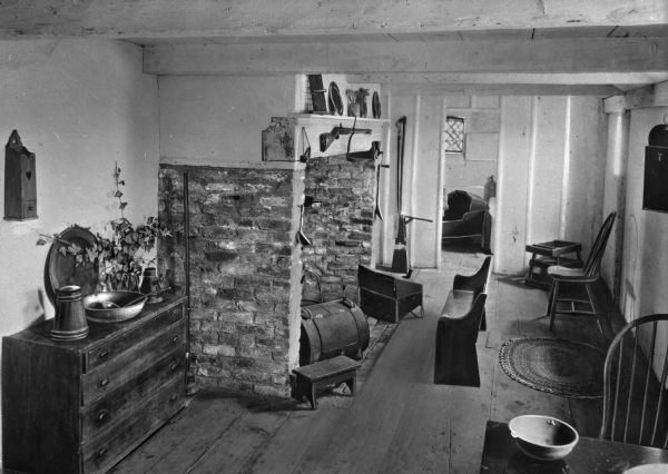 View of kitchen in the Jethro Coffin home. Jethro Coffin was a blacksmith and the grandson of one of the island's first white settlers, Tristam Coffin. Built in 1686 the house remains the oldest house on Nantucket. Room features a fireplace with mantle, dresser, table, chairs, bench and other wooden furniture. There is a small smoker in the fireplace, and a rifle above the mantle. In the next room is a wooden cradle.