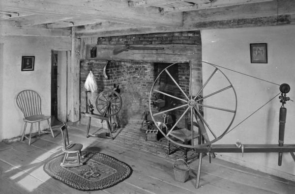 View of living room in Jethro Coffin home. Jethro Coffin was a blacksmith and the grandson of one of the island's first white settlers, Tristam Coffin. Built in 1686 the house remains the oldest house on Nantucket. The room features a brick fireplace, two spinning wheels, a small rug, a wooden chair, a Shaker-style chair and a child's chair. A rifle is above the fireplace, and there is a kettle in the fireplace as well as some other tools nearby.