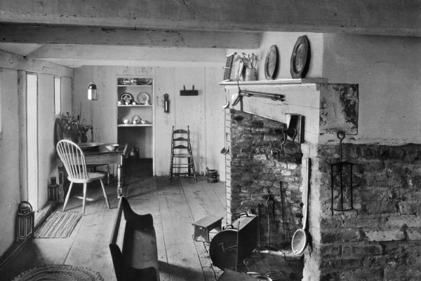 View of the kitchen and pantry in the Jethro Coffin home. Jethro Coffin was a blacksmith and the grandson of one of the island's first white settlers, Tristam Coffin. The house was built in 1686 and remains the oldest house on Nantucket. The room features a brick fireplace with a smoker and tools, mantle with a rifle and tableware, wooden bench, table and chairs. A pantry is in the background.
