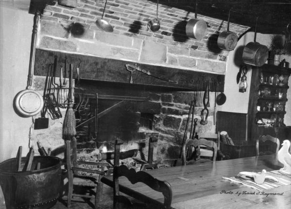 View of a seven foot tall colonial fireplace in a home. The large fireplace is surrounded by kitchen tools, fireplace tools and a bed warmer. A sword hangs over the fireplace, and  pots are hanging overhead. Elsewhere in the room are a kitchen table, chairs and a china cabinet. Carved wooden birds are on the table.