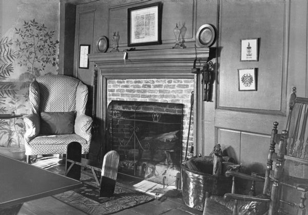 View of the parlor in the Denison homestead. Built in 1717, the home is a museum today. The room features a brick fireplace with mantle, a plush chair, two wooden chairs, a wooden bench and artwork. To the right of the fireplace a small wooden puppet hangs on the wall. A painted mural is on the adjacent wall.