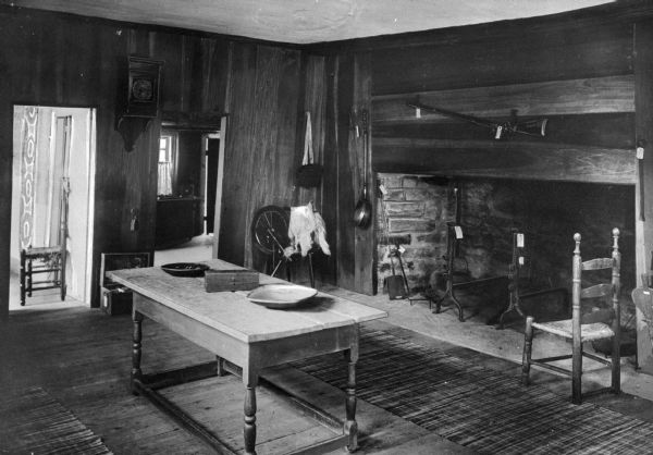 View of kitchen in Hyland House. Built in 1660, the house is still standing. Room features a fireplace with rifle hanging above, iron fireplace tools, a Shaker-style chair, table, spinning wheel and a clock.