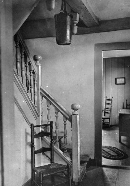 View of the stairway area in Hyland House. Built in 1660, the house is still standing. A Shaker-style chair is in the foreground next to the stairway, and another room is through a doorway.
