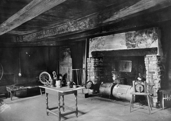 View of the lean-to kitchen in Hyland House, which was built in 1660. The room features a fireplace with two smokers, a table with several mortar and pestles, a spinning wheel, yarn swift and other household items. Wooden beams are on the ceiling.