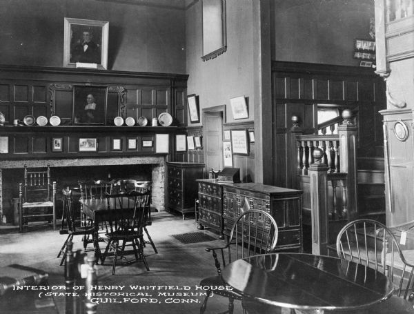 View of living room of the Henry Whitfield house. The room features a fireplace, tables, chairs, a mantle with tableware and framed portraits and artwork as well as other furniture. The house was built in 1639 and was the residence of Henry Whitfield, Guilford's first minister. Today it is the oldest remaining house in Connecticut and is operated as a museum. Caption reads: "Interior of Henry Whitfield House (State Historical Museum) Guilford, Conn."