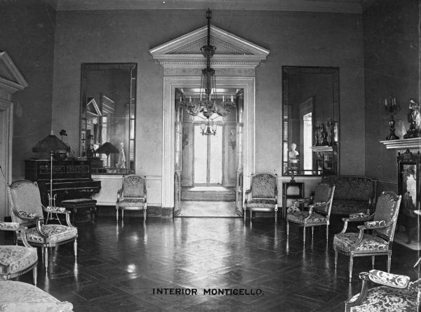 Interior view of Monticello, home of Thomas Jefferson. This view of the parlor displays Greek architectural decor and features a chandelier, large mirrors, sculpture, a fireplace, a small piano, chairs, a settee and a floor lamp. Caption reads: "Interior Monticello."