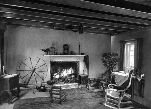 View of a living room in a colonial style home. Room features a fireplace, mantle with clock, cooking implements, candlesticks, a spinning wheel, tables and chairs and other decorative items.