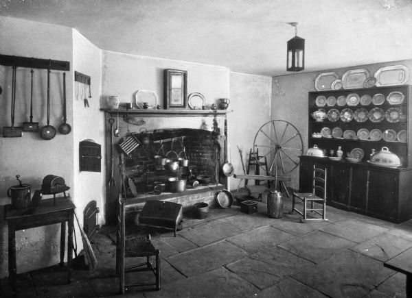 View of the old kitchen at the Telfair Academy. Room features a fireplace with cooking and other kitchen tools, a mantle with tableware and other small decorative items, a china cabinet displaying a large dish set, two chairs, a spinning wheel and various kitchen tools and implements. The Telfair Academy is one of the three buildings belonging to the Telfair Museum of Art.