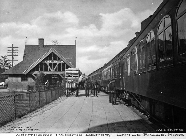 View of the Northern Pacific railroad depot. Men are standing next to the train parked in the station and the depot is on the left. "Little Falls" is the sign on the building. Caption reads: "Northern Pacific Depot, Little Falls, Minn."