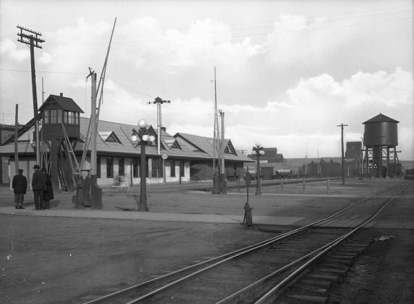 View of the Northern Pacific Railroad Depot, with three men standing in the empty train yard. In the distance is a water tower and other buildings.