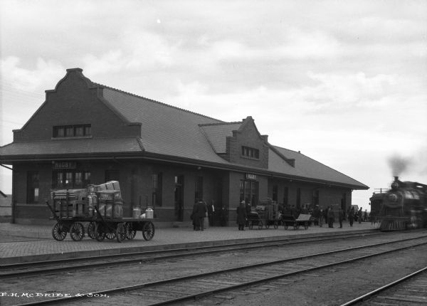 View of the Great Northern railroad depot, with a train pulling into the station while people wait near the depot. Several carts with luggage, milk containers and packages also await the train.