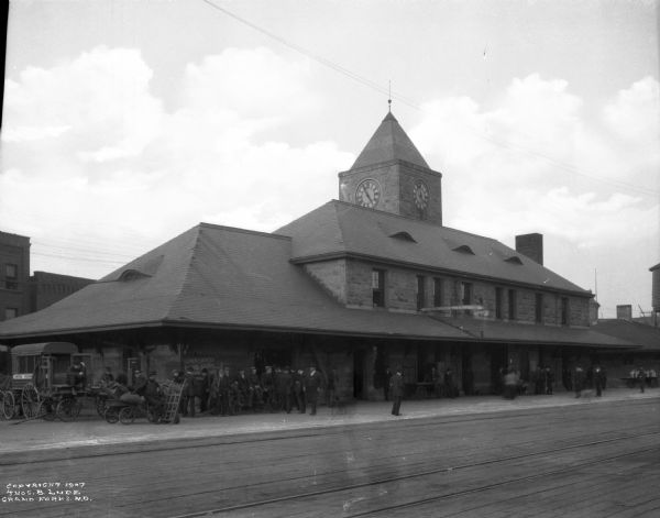 View of the Great Northern railroad depot. A group of men are in front of the depot. The clock on the building reads 4:55 and a sign on the depot reads: "Lunch Counter News Stand" (the rest is obscured).