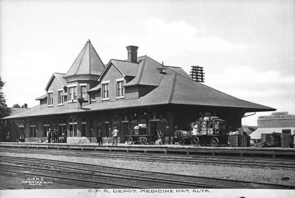 View of the Canadian Pacific Railway depot in Medicine Hat, Alberta, Canada. A number of men are waiting near the depot and on the right is a luggage cart loaded with bags. A sign on the building in the background reads: "Medicine Hat Dining Hall & Lunch Counter." Caption reads: "C.P.R. Depot, Medicine Hat, Alta."
