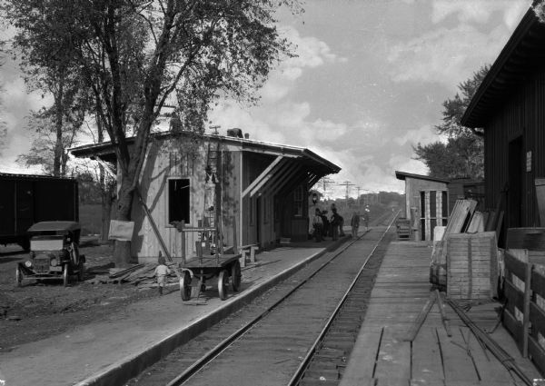 Several men are working on the railroad depot in Camp Dix, which was operated by Adams Express Company. Other men and a woman are standing near the building and a child is nearby. A sign on the cart reads: "Adams Express Company."