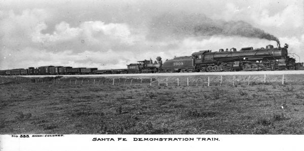 View of the Santa Fe demonstration train traveling through the countryside. A sign on the train reads: "A.T. & S.F." "3009" and "048." Caption reads: "Santa Fe Demonstration Train."