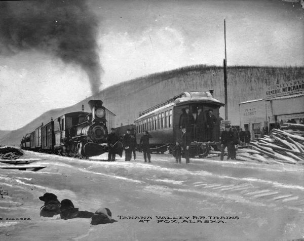 Several men and a child stand near a train equipped with a snow plow. A passenger car sits nearby, and three men, possibly railroad employees, stand on the outdoor deck of the car. Several dogs aren in the foreground. Signs on the building in the background reads: "P.C.S. Co. Market" and "Chelsey & Rya(obscured) General Merchandise Miners Supplies Steam Fitting." Caption reads: "Tanana Valley R.R. Trains At Fox, Alaska."