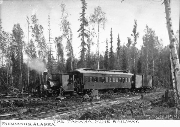 View of the Tanana Mine Railway train parked in a wooded area. Text on what appears to be a passenger car reads: "T.M.W." Caption reads: "Fairbanks, Alaska" and "The Tanana Mine Railway."