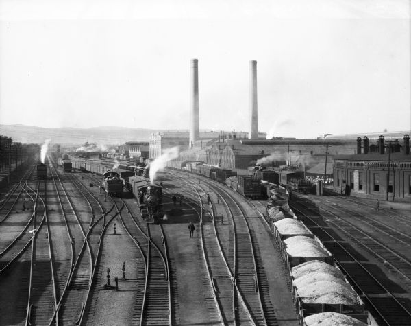 Elevated view of Lehigh Valley train yard with trains, cars full of goods, railroad workers and shops. One of the buildings features two large smokestacks.