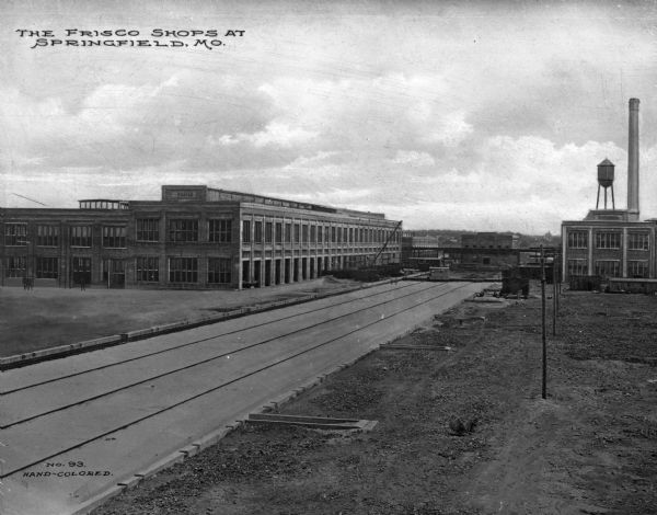 View of the St. Louis-San Francisco railway shops and train yards. The building on the right has a smokestack and what appears to be a small water tower. Several small cars, either railroad cars or trailers used in construction, are on the left side of the track area. Text on the cars reads: "Frisco."Caption reads: "The Frisco Shops At Springfield, Mo."