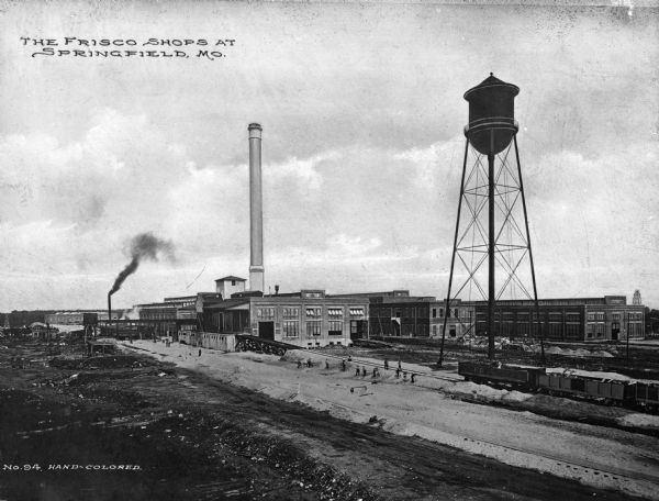 View of the St. Louis-San Francisco train yard and railway shops. Train cars are parked on the track leading into a warehouse. Men are working near the track and other warehouse style buildings are nearby, as well as a water tower. Text reads: "The Frisco Shops At Springfield, Mo."