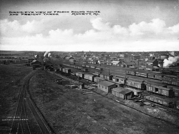 An elevated view of Frisco roundhouse and freight yards, a train yard where many cars and a few locomotives are parked. Painted on the side of many of the cars are signs that read: "Frisco." The town is on the right. Text at bottom reads: "Bird's-Eye View of Frisco Round House And Freight Yards, Monett, Mo."