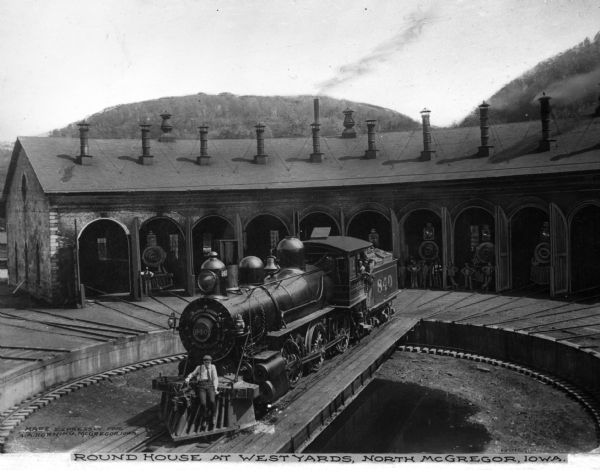 View of west yards train depot roundhouse. Railroad workers pose with locomotive engines in the roundhouse. The train in the foreground, no. "804," is on a turntable and a worker is posed on the front. Caption at bottom reads: "Round House At West Yards, North McGregor, Iowa." 