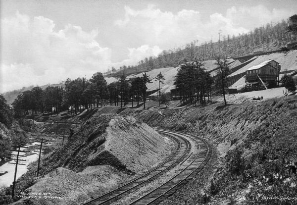Elevated view of the P & R coal storage yard, with railroad tracks winding around a curve at the foot of a small hill. The coal yard and buildings are at the top of the hill.