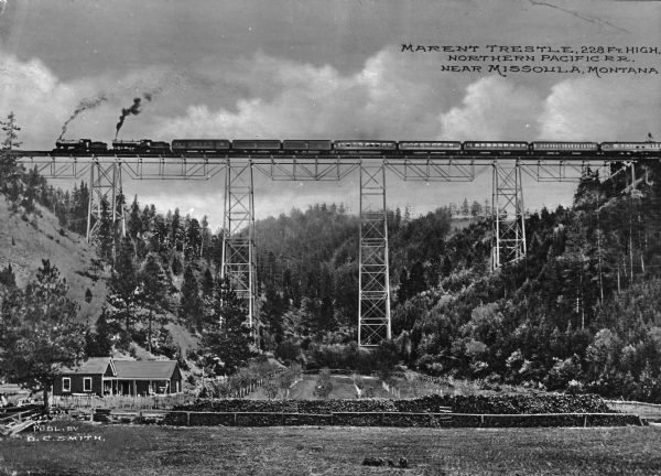 View looking up at the Marent trestle, part of the Northern Pacific Railroad. A train carrying passenger cars passes over the 228 foot high trestle, and a house and garden are far below. Caption reads: "Marent Trestle, 228 ft. high, Northern Pacific R.R. near Missoula, Montana."