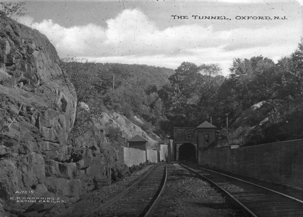 View of the entrance to a railroad tunnel which travels under a mountain. There are two small structures, one on the left of the railroad track and one on top of the opening. Caption reads: "The Tunnel, Oxford, N.J."