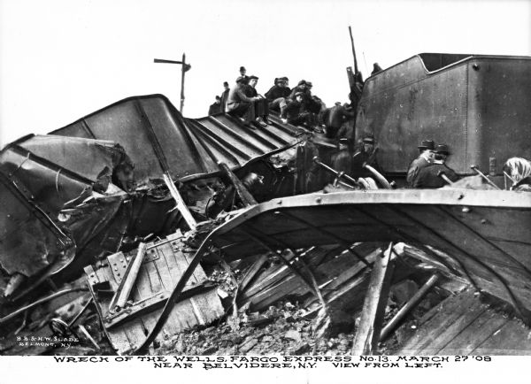View of the train wreck of the Wells Fargo Express, March 27th. Several men sit on the wreckage, and several other men and a women are nearby. Captionh reads: "Wreck of the Wells Fargo Express No. 13 March 27 '08 Near Belvidere, NY. View from left."