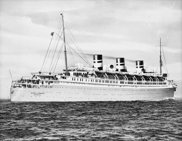 View of the "Queen of Bermuda" ocean liner in the water near New York City. The large ship features three large smokestacks, two masts, and many lifeboats. Passengers are on the decks. The sign painted on the side of the ship reads: "Queen of Bermuda Bermuda."