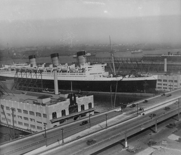 Elevated view of the "Queen Mary" ocean liner in port. The large ship features three large smokestacks, two masts and lifeboats. "Queen ary" is painted on the side of the ship. A highway and other businesses aren nearby. The sign on the building next to the highway reads: "French Line To All Europe."