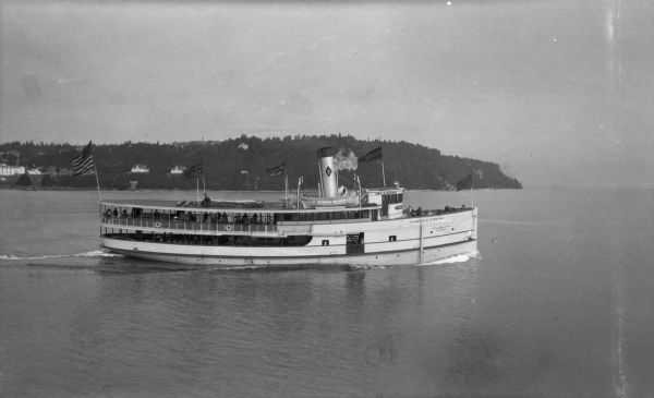 View of the steamer ship "Islander" leaving Mackinac Island. Signs painted on the ship reads=: "Islander," "Arnold Line" and "St. Ignace Mackinac Island" (bottom part of text illegible). Several flags fly on the ship, including an American flag and flags advertising the ship's destinations. Passengers are on the ship's decks.