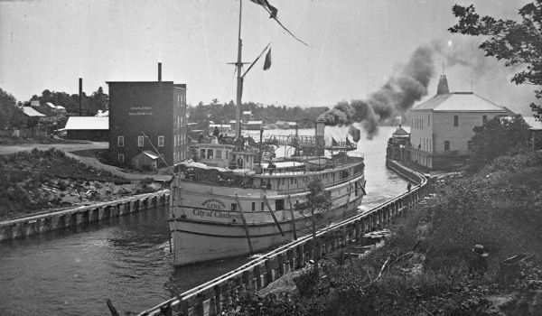 Elevated view of the "City of Charlevoix" ship coming into harbor. The sign painted on the front of the ship reads: "Northern Michigan Line" and "City of Charlevoix." People are on the boat's decks. In the background is a building with a sign reading: "Charlevoix Roller Mills."