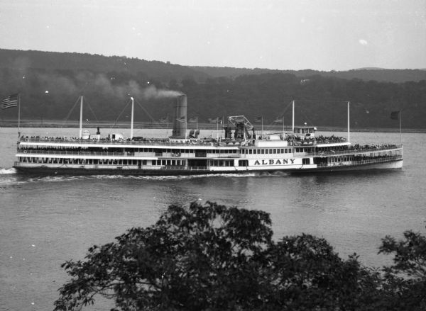 Elevated view over tree tops of the "Albany" steamship on the Hudson River near West Point. The large ship features three decks full of passengers, several American flags, four masts and a large smokestack. "Albany" is painted on the side of the vessel.