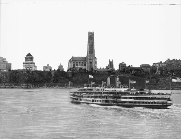 View across water toward the "Hendrick Hudson" excursion boat near New York City. Many passengers are on the large excursion boat's three decks. The boat also features several American flags and two smokestacks. "Hendrick Hudson" is painted on the side of the ship. Some of the city's buildings, including a large church, are in the background.
