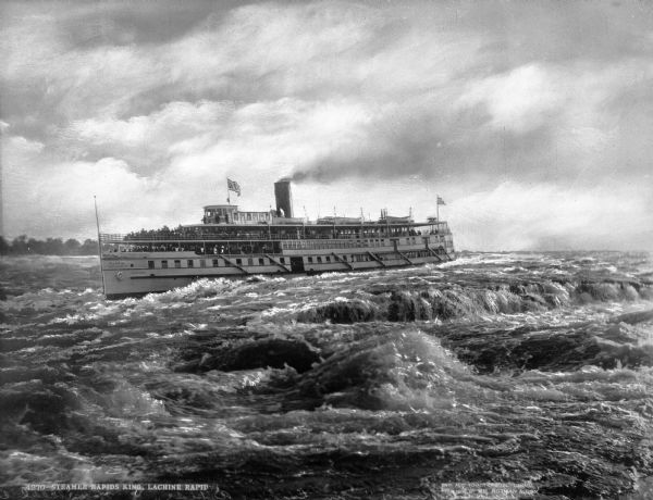 View of the steamship "Rapid King" in the Lachine Rapids in the Saint Lawrence river near Montreal, Quebec, Canada. Many passengers are on board the ship, which is making its way through the rapids. The ship features one smokestack, lifeboats and two flags. Signs painted on the boat read: "R. & O.N. Co." (rest of text illegible) and "Rapid King." Caption reads: "4270- Steamer Rapids King, Lachine Rapid."