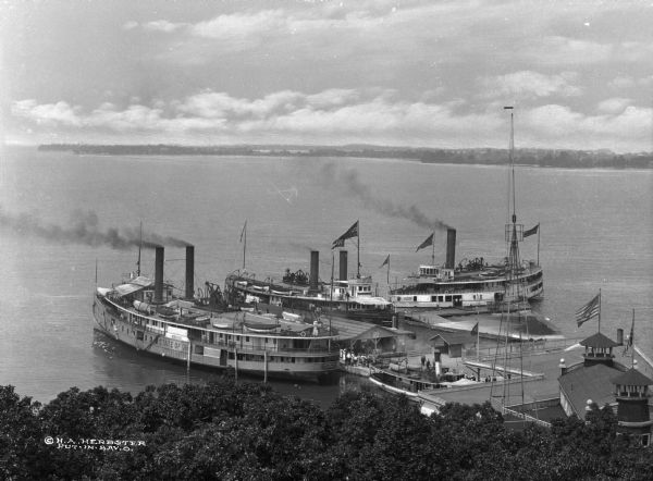 Elevated view of three steamships arriving at Fox's Dock, an island in Lake Erie. People are waiting on the docks. A smaller ship is in the dock area as well. The ship on the left side of the dock reads: "State of Ohio" on its side and "State of Ohio of Cleveland" on its front. The ship on the right side of the dock has a name that reads: "State of New York" and "Toledo, Put-In-Bay & Cleveland." The third ship, on the far right, has a painted sign that reads: "Put-In-Bay Route" "U.S. Mail" and "Frank E. Kirby."