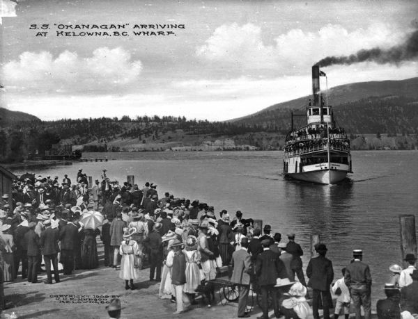 View of the steamship "Okanagan" arriving in Kelowna, British Columbia, Canada. Passengers are on board the ship, and a crowd awaits the ship's arrival on the dock. Caption reads: "S.S. 'Okanagan' arriving at Kelowna, B.C. Wharf."