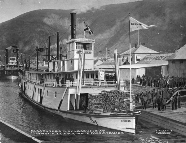 View of passengers disembarking from a White Pass steamer ship in Dawson, Yukon Territory, Canada. The ship is marked: "Dawson." Piled wood is on the front of the ship, and several flags fly, one reading: "Royal" (rest of text obscured). A crowd is standing on the dock. Signs for "White Pass &" (obscured) are on the nearby building. A mountain is in the background. Caption reads: "Passengers disembarking at Dawson, Y.T. from White Pass Steamer."