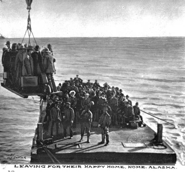 View of passengers disembarking from a lighter by means of a platform suspended from a derrick in the water near Nome. Text on photograph reads: "Leaving for their happy home, Nome, Alaska" and "By O.D. Goetze."