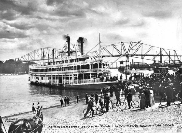View from shoreline towards a docked Mississippi riverboat. Passengers are disembarking from the ship, and a large crowd is gathered on shore, with some people on bicycles. A bridge is in the background. Caption reads: "Mississippi river boat landing Clinton, Iowa."