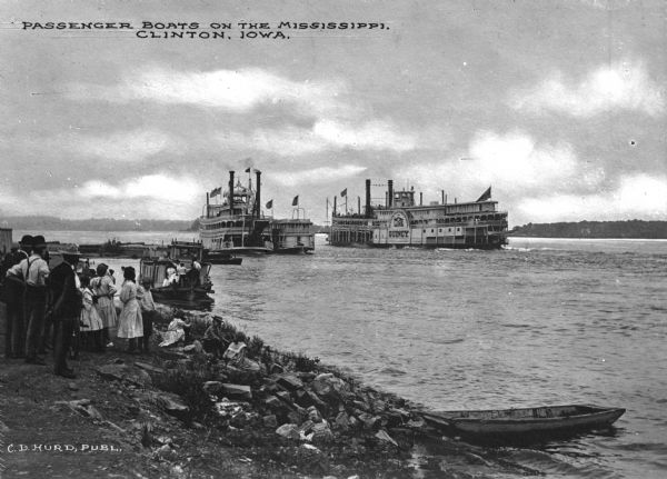 View from shoreline of two passenger steamboats on the Mississippi river near Clinton. Several children and adults wait on shore. A tugboat is nearby in the river, and another small vessel is near one of the steamboats. The sign painted on one of the ships reads: "Quincy Line St. Louis Davenport St. Paul." Caption reads: "Passenger boats on the Mississippi. Clinton, Iowa."