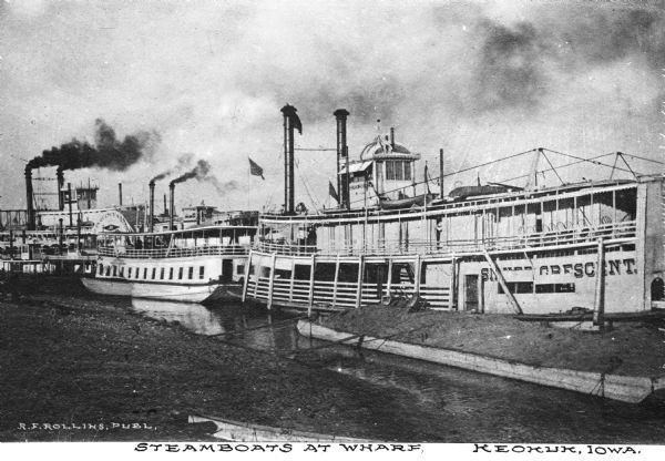 View of several steamboats docked at the wharf. The ship on the right is the "Silver Crescent." The ship on the far left bears the logo "St. Louis Quincy &" (rest is illegible). Caption reads: "Steamboats At Wharf Keokuk Iowa."