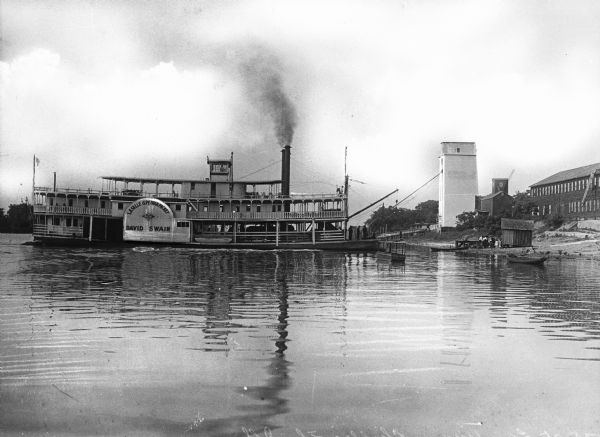 View of the steamship "David Swain" docked in the water near Chillicothe. The sign painted on the side of the ship reads: "Lasalle & Peoria Packet David Swain" and "The Royal" (rest of logo illegible). People are on board the boat, and also waiting on the shore. A large warehouse and other buildings are on shore.