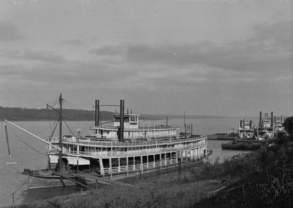 View of several boats docked along the shore. The sign painted on the boat on the far left reads: "Rees Lee." One of the boats on the right bears the name "Barrett" and the letter "B."