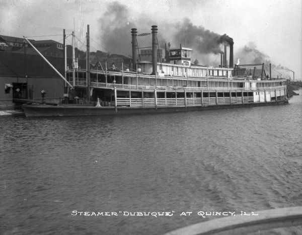 View of the steamboat "Dubuque" docked in the Mississippi River. Passengers are on the ship decks and smoke is coming out of several nearby smokestacks. Caption reads: "Steamer 'Dubuque' at Quincy, Ill."