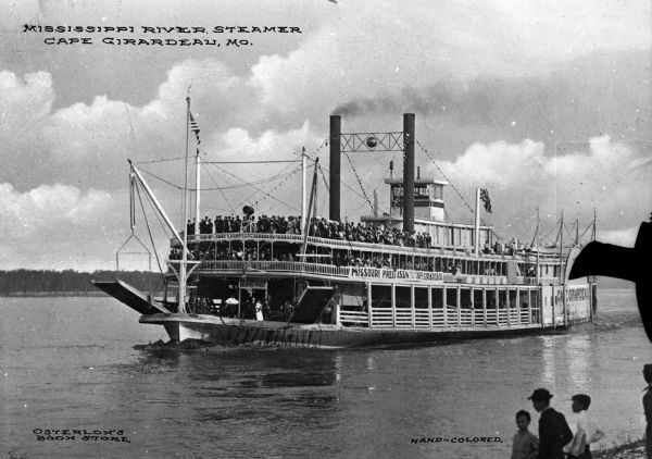 View from shoreline of the steamship "Cape Girardeau" in the Mississippi River near Cape Girardeau. Many passengers are on board the ship as it prepares to land. Several boys are waiting on shore. Sign on the front of the ship, which is partially obscured, reads: "Imperial Club Welcomes The Press." Sign on the side of the ship reads: "Missouri Press Assn" and "Cape Girardeau." "Cape Girardeau" is painted on the rear of the ship. Text on the photograph reads: "Mississippi River Steamer Cape Girardeau, Mo."