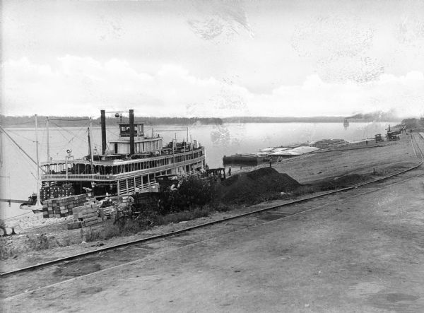Elevated view of the steamboat "Bald Eagle" unloading on the riverfront. "Bald Eagle" is painted on the top of the ship. Several men are working on and near the boat. Cargo is piled on shore and on the ship. Another steamboat is in the distance.